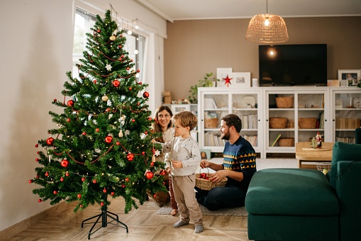Young family is sitting in front of christmas tree and decorating it. They are wearing winter sweaters. They are smiling and enjoying each others company. Horizontal photo with copy space. Bright and cozy living room atmosphere.