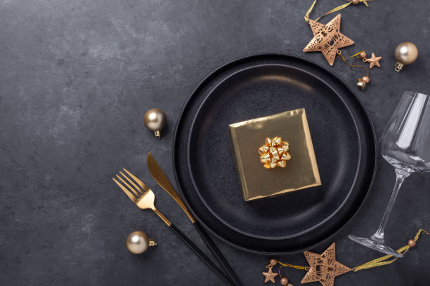 istock Christmas table setting with black ceramic plate, glass, gift box and gold accessories on black stone background. Top view. Copy space 1279335078