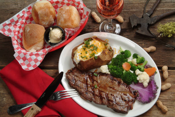 A Porterhouse steak dinner with a Western motif. A white platter with a BBQ Porterhouse steak with garnished baked potato and steamed vegetables. along side is a basket of dinner rolls, and a glass of rose wine served on a barn wood table adorned with a pair of fence pliers and moss covered wood