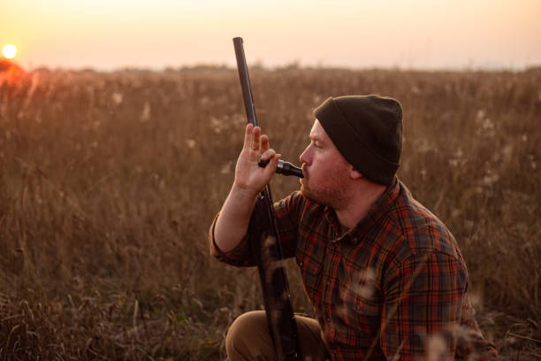 Young red bearded hunter  sitting it grass and blowing at hunter's whistle - photo with selective focus on his hand