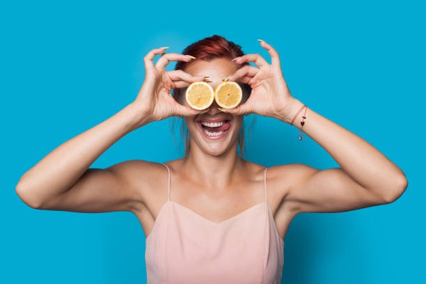 Red haired fit woman covering her eyes with a sliced lemon is smiling on a blue studio wall Red haired fit woman covering her eyes with a sliced lemon is smiling on a blue studio wall woman applying wallcovering stock pictures, royalty-free photos & images