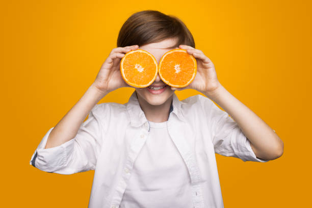 istock Cute boy covering his eyes with sliced oranges and smile on a yellow studio wall 1279353786