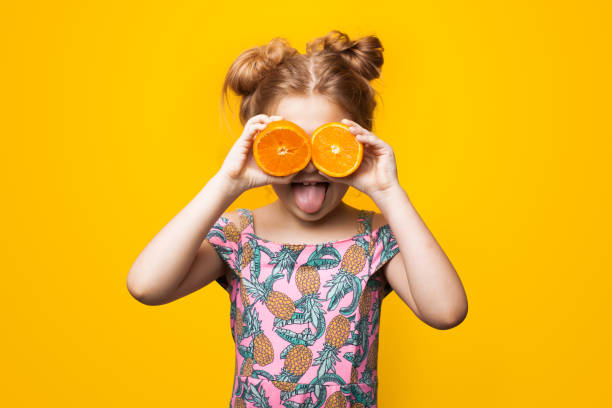 petite fille caucasienne avec les cheveux blonds et la robe d’été pose avec des oranges tranchées sur des yeux et la bouche ouverte sur un mur jaune de studio - isolated on white fun orange food photos et images de collection