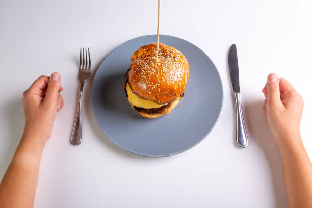 Homemade cheeseburger on a gray plate and a person with cutlery in his hands on a white background. Hands in the frame. View from above. Horizontal orientation. High quality photo