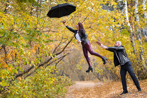 Autumn. A beautiful dark-haired girl flies away on an umbrella from a bald brutal guy. Girl flying on an umbrella.  A man and a woman in the autumn forest. Horizontal photography