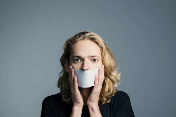 beau jeune homme avec de longs cheveux blonds a couvert sa bouche avec une feuille blanche sur un fond gris. - censorship photos et images de collection