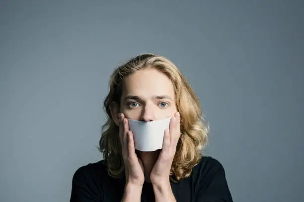 Photo of Handsome young man with long blonde hair covered his mouth with a white sheet on a gray background.