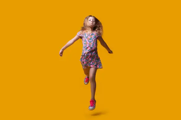 Photo of Cute blonde lady running on a yellow studio wall wearing a summer dress and gesturing a hurry