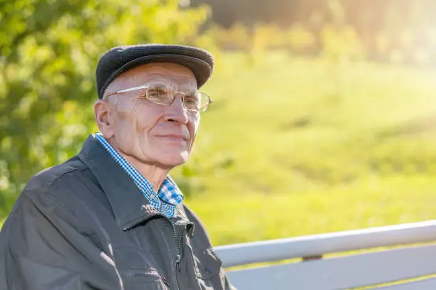 retired old mature citizen wearing cap and eyeglasses sitting on wooden bench and looking to side with smile on face outdoor