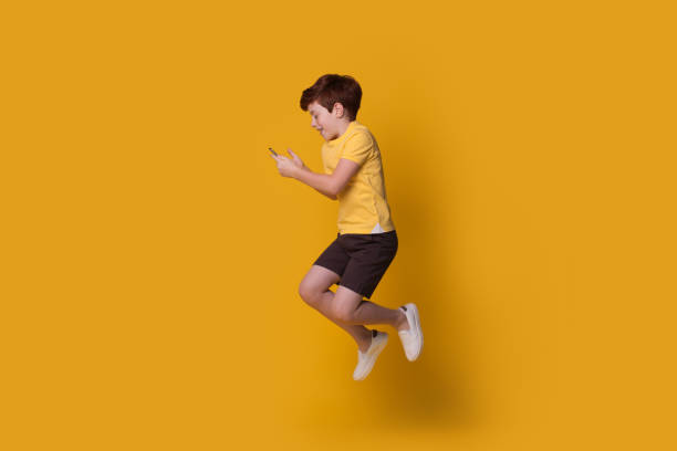 istock Ginger caucasian boy wearing a yellow t-shirt is jumping on studio wall while chatting on mobile 1279369983