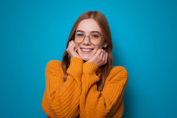 istock Charming ginger woman with glasses and yellow sweater is gesturing warmth on a blue studio wall smiling at camera through eyeglasses 1279375069