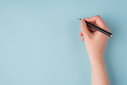 Top above overhead close up first person view photo of girl's right hand holding black pen starting to write isolated over blue color pastel background