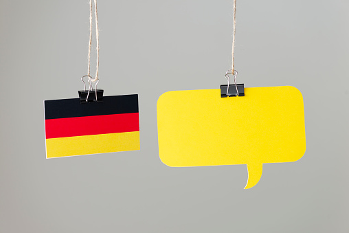 One yellow speech bubble and a German flag are hanging via ropes in front of gray background.