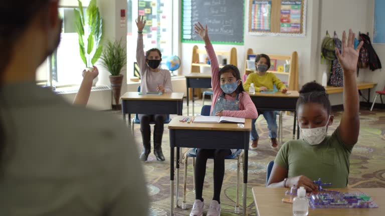 Group of elementary school classmates wearing protective face masks in the classroom while desks are socially distanced due to new COVID-19 regulations.