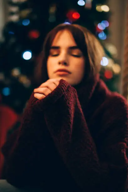 Pretty girl praying for Christmas. Dressed in an oversized sweater. Christmas tree behind the back.