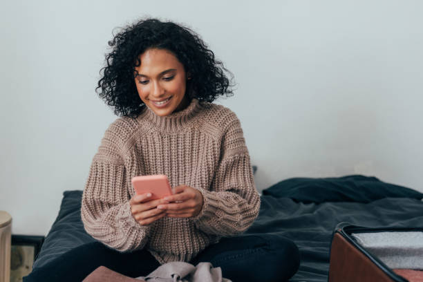 felice giovane donna imballaggio sua valigia per le vacanze invernali e la prenotazione di un volo online sul suo telefono cellulare - hotel wireless technology bedroom hotel room foto e immagini stock