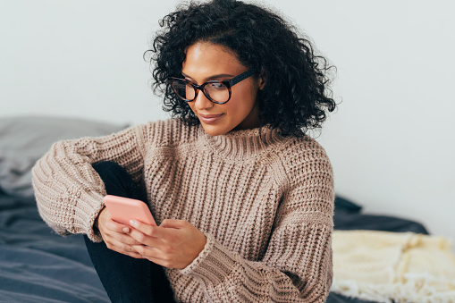 Beautiful young woman in knitwear sitting on her bed and texting on her smartphone.