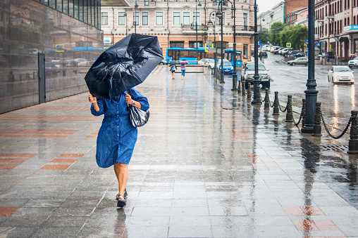 Vladivostok, Russia - August 6, 2020: Woman in bad rainy weather walks down the street and tries to keep the umbrella from the strong wind. City landscape in rainy weather.