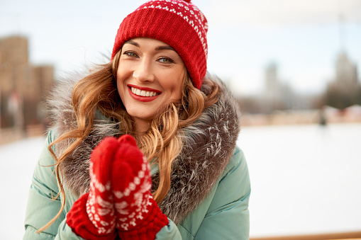 Beautiful lovely middle-aged girl curly hair warm winter jackets red knitted hat glove stands ice rink background Town Square Christmas mood lifestyle Happy holiday woman  snowy day Winter leisure