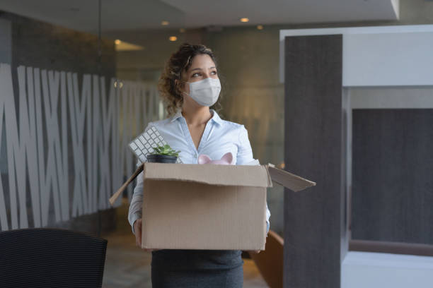 Worker wearing a facemask at the office while holding a box with her belongings after being fired Female worker wearing a facemask at the office while holding a box with her belongings after being fired from her job during the COVID-19 pandemic quitting a job stock pictures, royalty-free photos & images