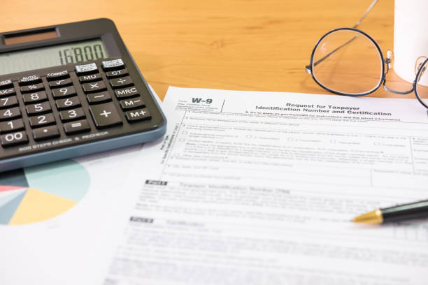 istock Selective focus Calculator button. American US tax form on wooden table. With blurred calculator, pen and glasses. Taxation time obligation for people who has income that need to comply to regulation. 1279436638