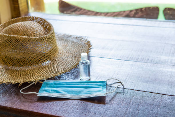 shot of surgical face mask, together with straw hat and on wooden table inside of the hotel. New normal lifestyle for traveler after Coronavirus. shot of surgical face mask, together with straw hat and on wooden table inside of the hotel. New normal lifestyle for traveler after Coronavirus. vacation rental mask stock pictures, royalty-free photos & images