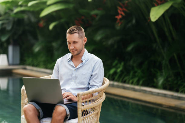 istock Young man using a laptop computer in a garden with a swimming pool. 1279441675
