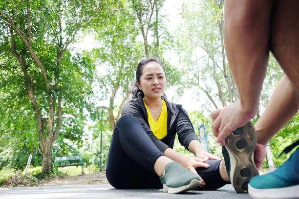 la giovane donna asiatica in abbigliamento sportivo è dolorosa e crampi alla gamba o alla caviglia e fidanzato che aiutano a rilassarsi della contrazione resiliente e muscolare dopo l'esercizio fisico e la corsa in giardino. - cramping foto e immagini stock