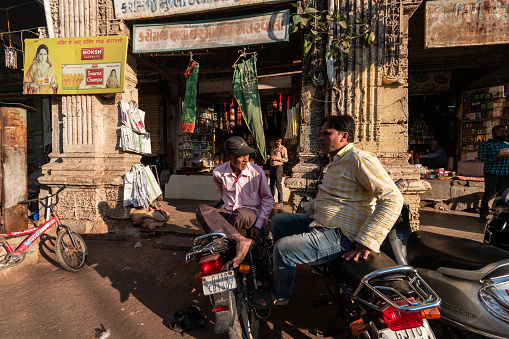 Jamnagar, Gujarat, India - December 2018: Two Indian men having a conversation sitting on their parked motorbikes in a market in the old city.