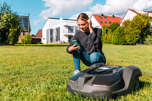 Young woman controlling a robot lawn mower with her smart phone in the summer. The woman kneeling next to the robotic mower, looking to her smart phone to program the robot mower. Modern Home in the background.