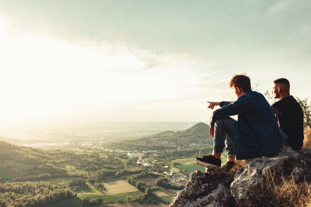 Two male friends sitting together side by side on a mountain top rock, enjoying the sunset view in the middle of the green outdoor nature. One man pointing to the valley village, explaining his friend the sunset valley view. Millennial Generation - Youth Culture Best Friends Outdoors Lifestyle Concept.