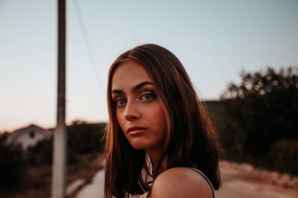 A girl in a denim suit with a backpack walks in the center of a quiet road among the mountains at sunset