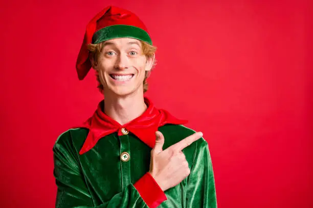 Photo of positive elf guy point finger empty space x-mas christmas ads wear costume cap isolated on bright red color background.