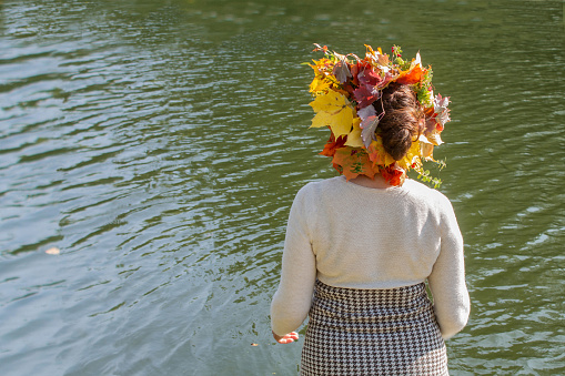 A woman in a wreath of autumn leaves, in a light-colored dress by the water, looks into the distance. Autumn wreath - a symbol of the successful end of the agricultural season, a rich harvest and femininity