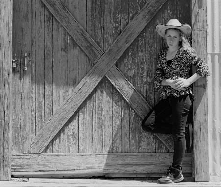 Cowgirl leaning on rustic barn door, copy space, black & white. Horizontal.
