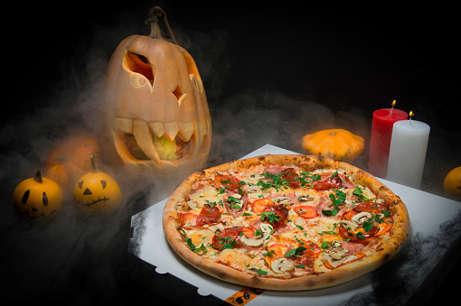 meat pizza on delivery box near Halloween decoration. Scary pumpkins with painted faces, Lights of burning candles In horror smoke. Dark background with cobwebs and spiders near fast food