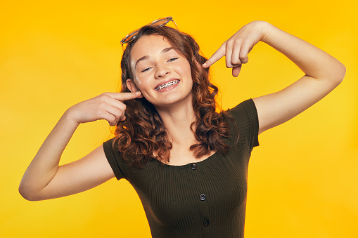 Young woman teen girl smiling pointing with fingers on teeth with braces, on yellow background with glasses in her hair