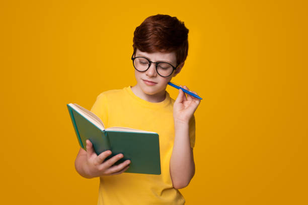 istock Concentrated caucasian boy is reading a book on a yellow studio wall holding a pen and wearing glasses 1279507729