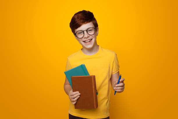 istock Young ginger schoolboy holding some folders is smiling on a yellow studio wall while wearing glasses and casual t-shirt 1279519808