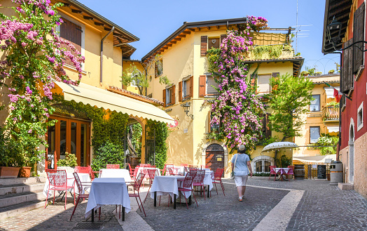 Gardone Riviera, Lake Garda, Italy - September 2018: Person walking past a restaurant with outside tables in Gardone Riviera. The buildings are covered by large flowering climbing plants.