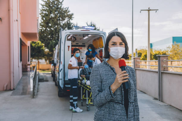 Newscaster woman with face mask reports breaking news in front of hospital during pandemic Journalist and TV news reporter young woman wearing a prevention facemask speaking and presenting the breaking news into a microphone during broadcast and making reportage about a Covid-19 virus epidemic in front of a hospital and ambulance staff. Interviewed people during coronavirus quarantine and lockdown. tv reporter photos stock pictures, royalty-free photos & images