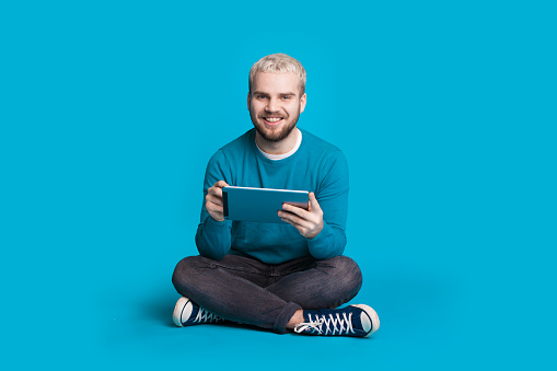 Blonde man with beard is using a tablet while sitting on floor at a blue studio wall smiling at camera