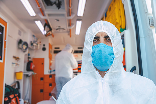 Portrait of first aider healthcare emergency staff young doctor man dressed white protective medical disposable hooded coverall suit with glove and face shield and N95 mask gear before the decontamination process for EMT, standing, thumbs up, smiling, looking at the camera posing in front of ambulance car during the covid19 pandemic after coronavirus quarantine and lockdown