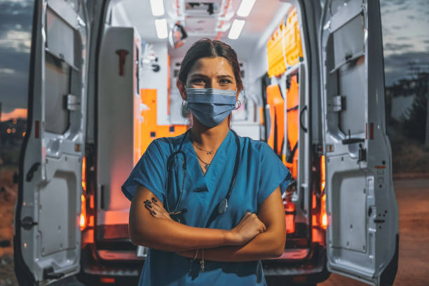 Portrait of young female paramedic with face mask working in an ambulance during pandemic Portrait of a paramedic healthcare worker nurse woman wearing a surgical face mask working night shifts for EMT, standing, arms folded, smiling, looking at the camera posing in front of ambulance car during the covid19 pandemic after coronavirus quarantine and lockdown frontline worker mask stock pictures, royalty-free photos & images