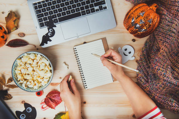 Top view woman writing plans or list of films for Halloween movie party. Blank notebook, laptop, popcorn bowl, pumpkin, bat, ghost, fall leaves, warm plaid on wooden background. Cozy and safe holiday top ver la mujer escribir planes o lista de películas para la fiesta de cine de halloween. cuaderno en blanco, computadora portátil, cuenco de palomitas de maíz, calabaza, murciélago, fantasma, hojas de otoño, a cuadros calientes sobre fondo de madera