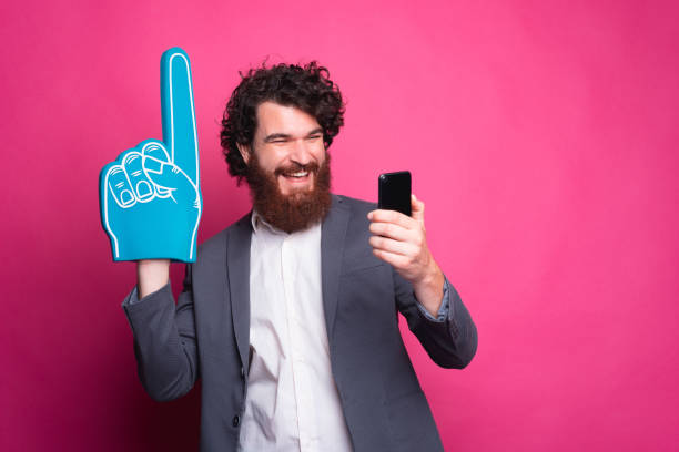 istock Happy man supporting his favorite team, excited bearded man in casual using phone and pointing with fan glove 1279546996