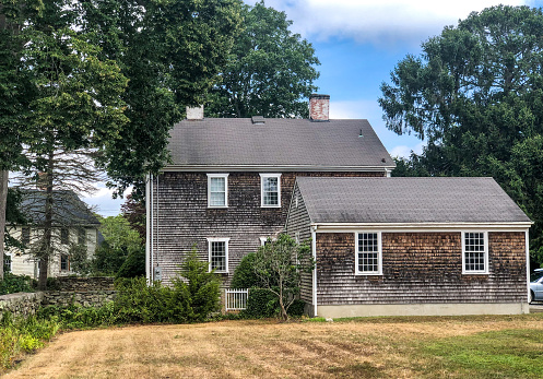 Adamsville, RI USA - August 2, 2020: A side view of an old Colonial house and garage with weathered shake shingles in a New England village near the ocean. A stone wall on the left surrounds the yard and the dry grass.