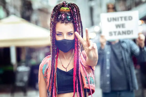 Young mixed race woman wearing face mask showing peace sign during street protest – Hipster female protesting outdoor for human rights with people holding sign board behind her in city