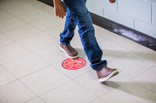 Cropped view of an elementary student walking in a school hallway. He has returned to the classroom during the covid-19 pandemic. Stickers on the floor are used to keep the children six feet apart according to social distancing guidelines.