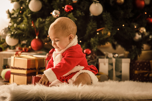 Full length shot of happy little baby girl in a festive outfit sitting on the cozy white faux fur rug next to the Christmas tree and having fun while playing with a gift box.